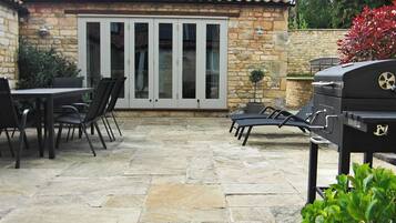 A patio with stone flooring, a table with chairs, a grill, and a stone wall with a window.