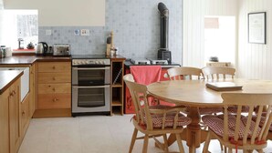 Une cuisine avec des meubles en bois, un four double, une cuisinière à gaz et un plan de travail carrelé. On y trouve une table à manger avec des chaises et une nappe rouge.