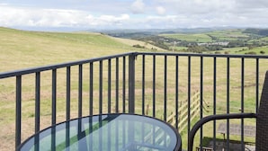 Un balcón con mesa y silla de vidrio, con vistas a un paisaje rural con colinas onduladas y un pequeño pueblo a lo lejos.
