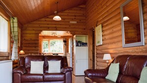 A cozy cabin interior with a brown leather sofa, a wooden dining table, and a kitchen area visible through a window. - Fingask Log Cabin - S4394 (Glenfarg)