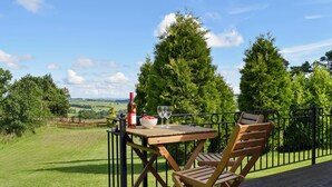 A wooden table with a bottle and glass on a balcony overlooking a grassy landscape with trees. - Fingask Log Cabin - S4394 (Glenfarg)