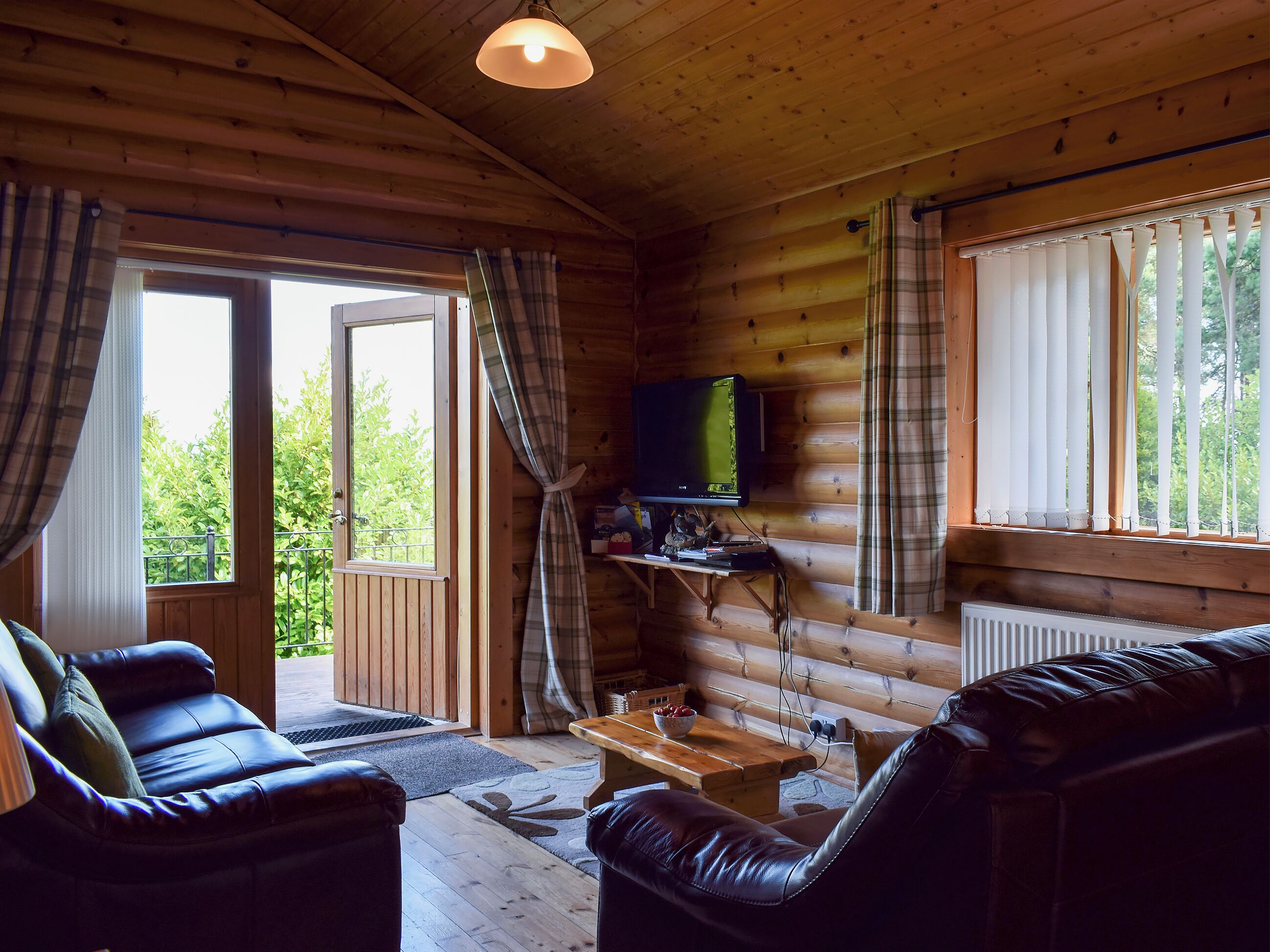 L’intérieur chaleureux d’un chalet, avec un revêtement mural en bois, un téléviseur posé sur une étagère et une grande fenêtre agrémentée de rideaux.