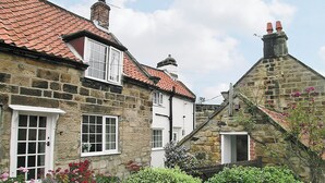 Un bâtiment traditionnel en pierre, avec un toit recouvert de tuiles rouges, des encadrements de fenêtres blancs et un petit jardin agrémenté de fleurs roses.