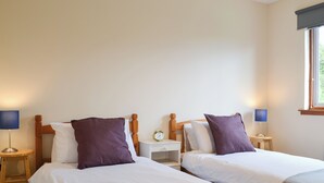 A hotel room with two single beds, each with a wooden headboard, white bedding, and a purple pillow. There are bedside tables with lamps, a small clock, and a window with a view of greenery. - 2 bedroom accommodation in Ardentinny, near Dunoon (Ardentinny, near Dunoon)