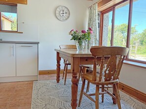 A dining area with a wooden table, chairs, a vase with flowers, a wall clock, and a window with a view of greenery. - 2 bedroom accommodation in Near Marazion (Near Marazion)