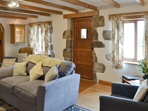 Un salon chaleureux avec un canapé, des fauteuils, une porte en bois, des murs en pierre et une fenêtre ornée de rideaux à motifs floraux.