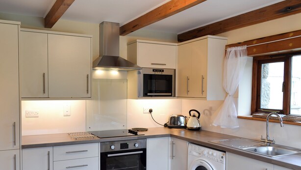 Cocina compacta con horno empotrado, microondas y lavadora, vigas de madera y una ventana con cortina.