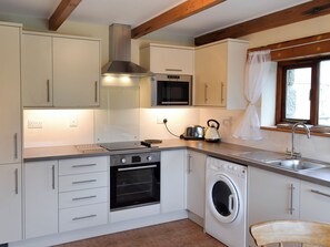 Une cuisine compacte dotée d’un four, d’un micro-ondes et d’un lave-linge encastrés, de poutres en bois et d’une fenêtre avec un rideau.