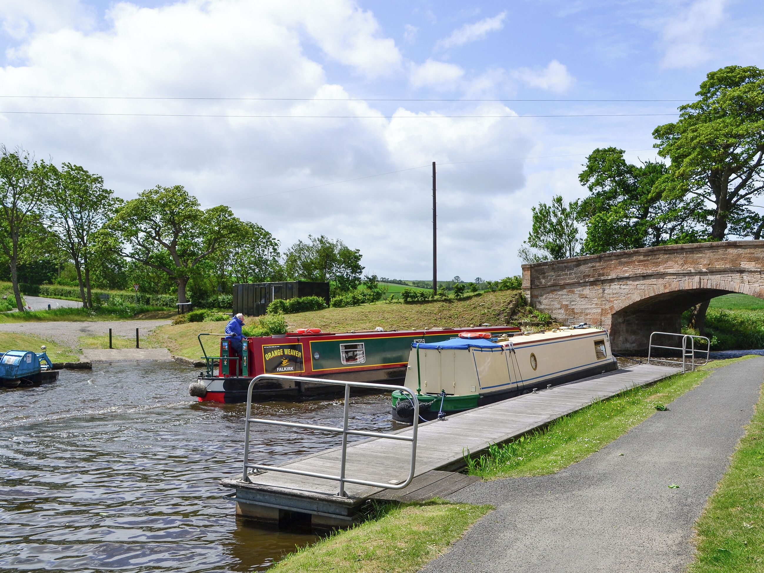 Un canal estrecho con un barco fluvial amarrado en una esclusa, un puente de piedra y una orilla cubierta de césped.