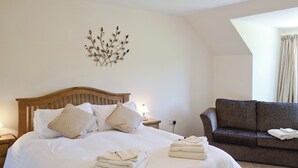 Un lit bien fait, avec du linge de lit blanc et deux serviettes pliées, une tête de lit en bois, une décoration murale en forme de feuille, un canapé marron avec une serviette blanche pliée, et une fenêtre avec des rideaux.