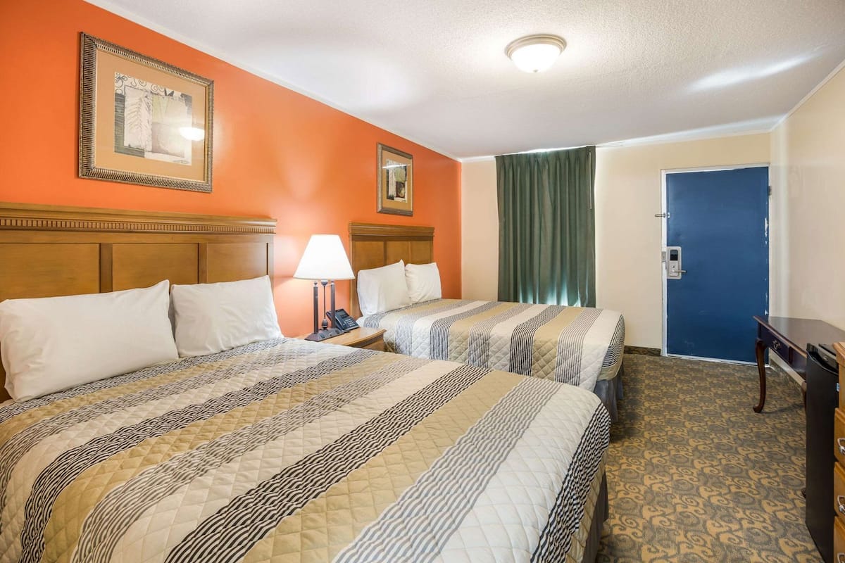 A hotel room with two beds, a nightstand with a lamp, a blue door, and a patterned carpet.