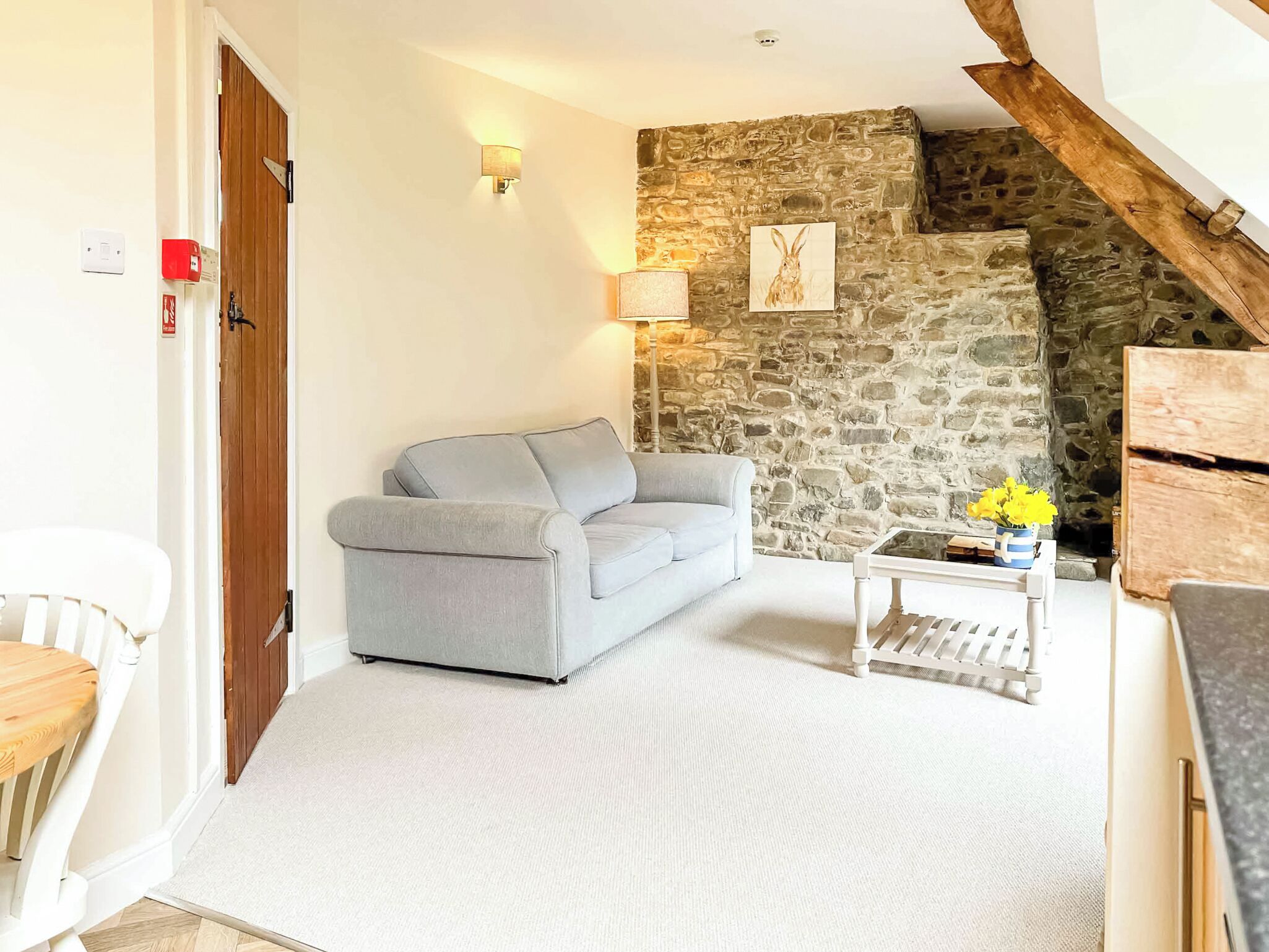 Ein Wohnzimmer mit grauem Sofa, einem kleinen Tisch mit Blumen, einer Steinwand und einer Holztür.