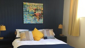 Ein ordentlich bezogenes Bett mit blauem Kopfteil, zwei Nachttischlampen und einem Gemälde mit Booten an der Wand.