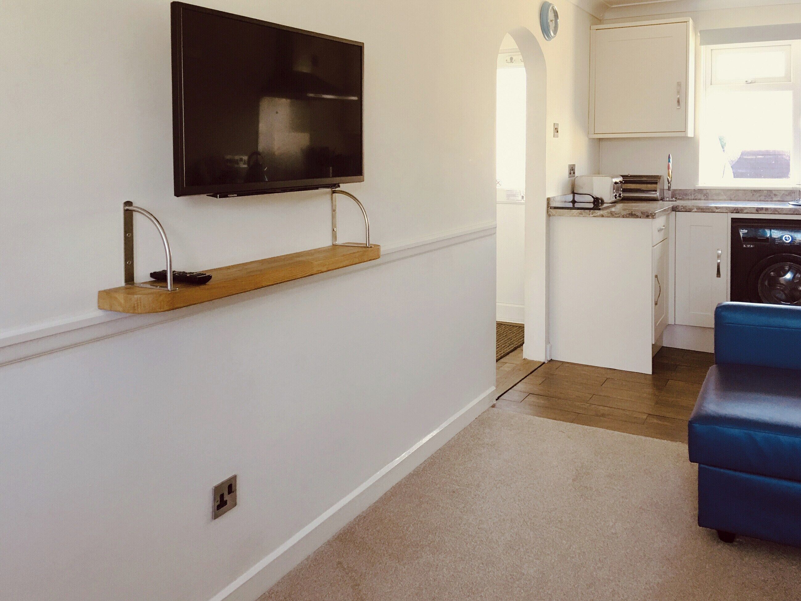A compact kitchen with a built-in dishwasher, a flat-screen TV mounted on the wall, and a wooden shelf.