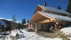 A wooden lodge with a snow-covered roof, a wooden porch, and a mountain backdrop. - Familiy friendly holiday apartement in a ski area (Sirnitz)