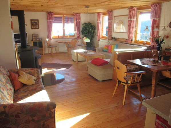 Una sala de estar acogedora con sofá, reposapiés y sillones, suelos de madera y ventanas grandes con cortinas a cuadros.
