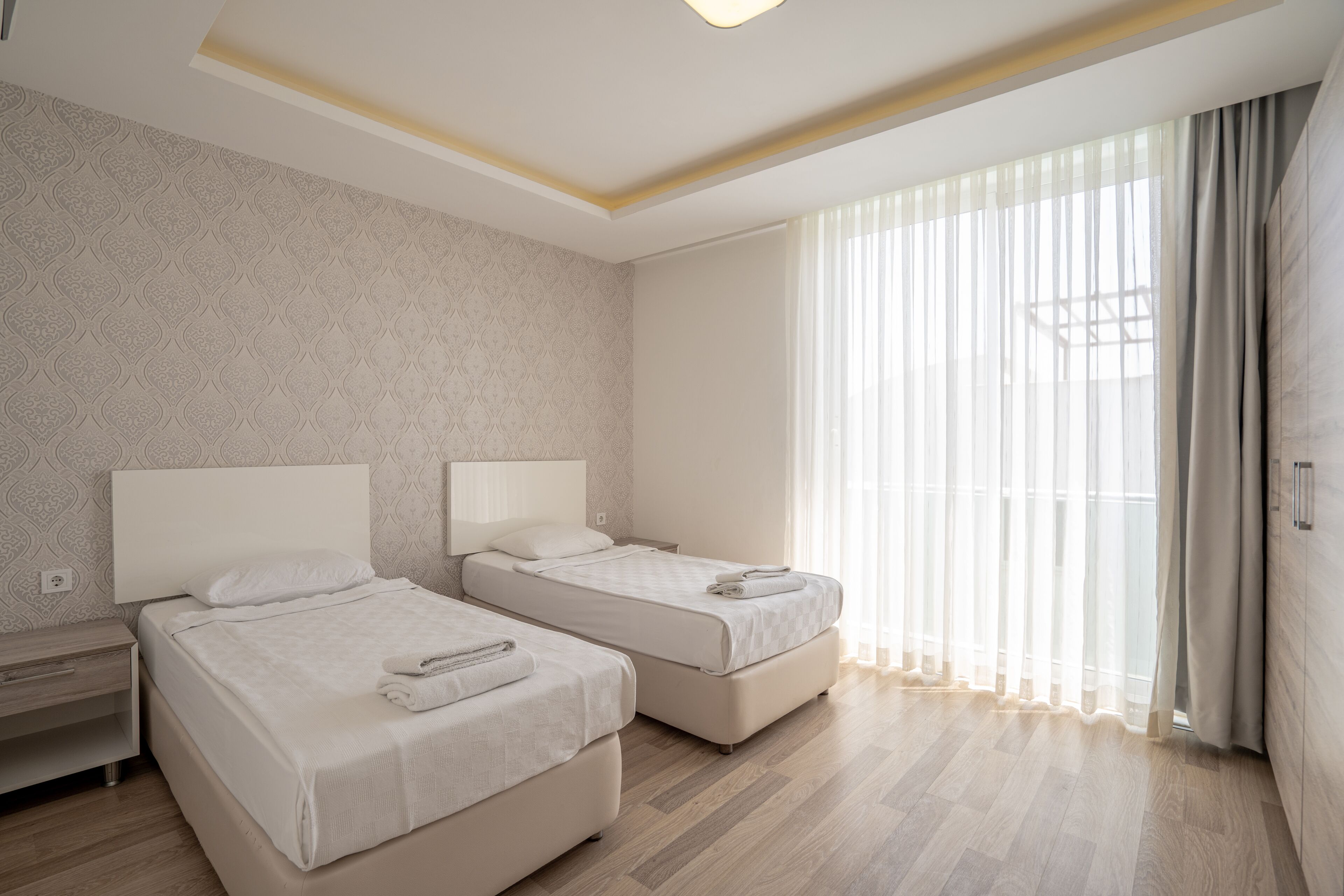 A hotel room with two beds, a wooden floor, a patterned wall, and a large window with curtains.