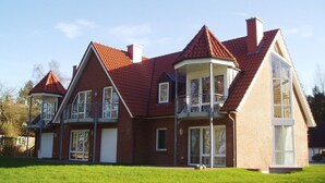 Ein zweistöckiges Haus mit rotem Ziegeldach und einem Balkon im zweiten Obergeschoss sowie einem großen Fenster im ersten Stock.