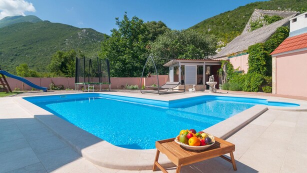 Une piscine avec une table en bois sur laquelle sont disposés des fruits, le tout entouré d’une clôture, avec une montagne en arrière-plan.