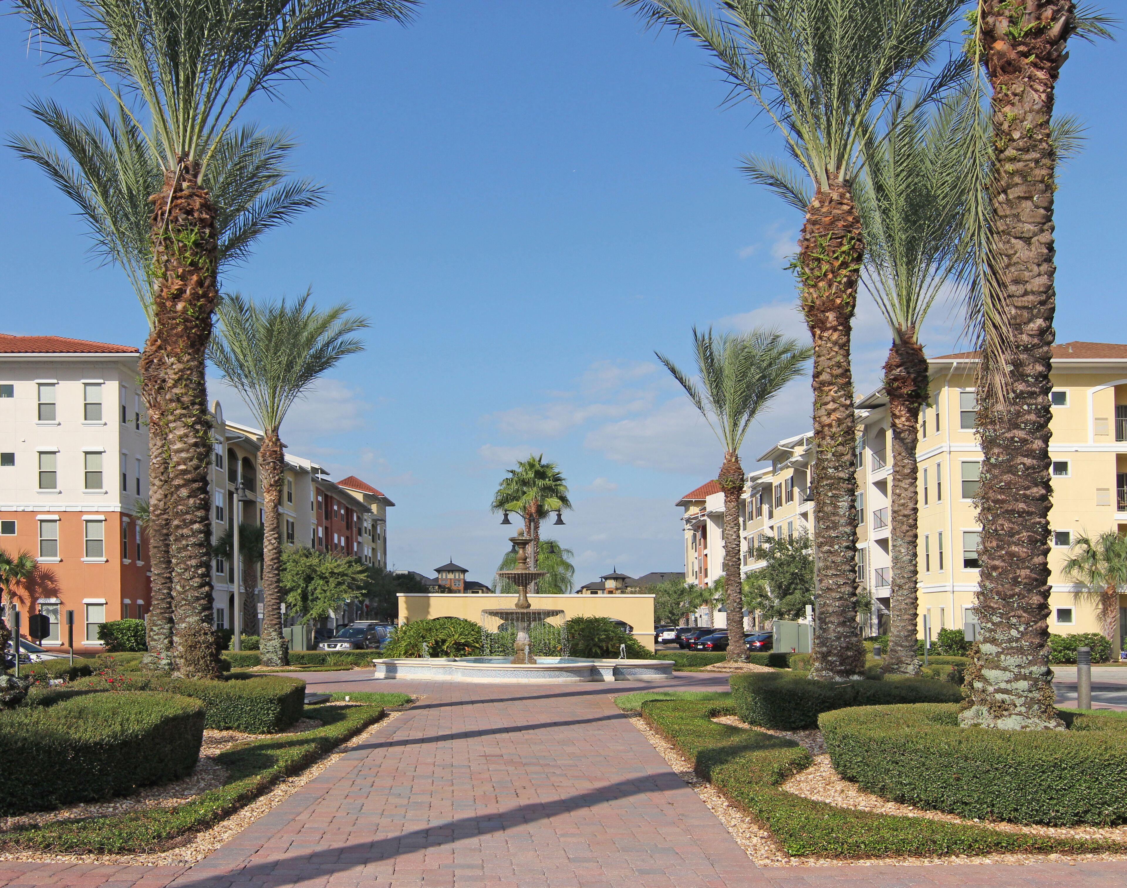 A pathway flanked by palm trees leading to a central fountain, surrounded by neatly trimmed hedges and multi-story buildings with balconies.