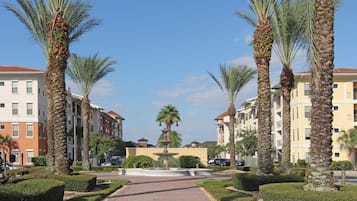 A pathway flanked by palm trees leading to a central fountain, surrounded by neatly trimmed hedges and multi-story buildings with balconies.