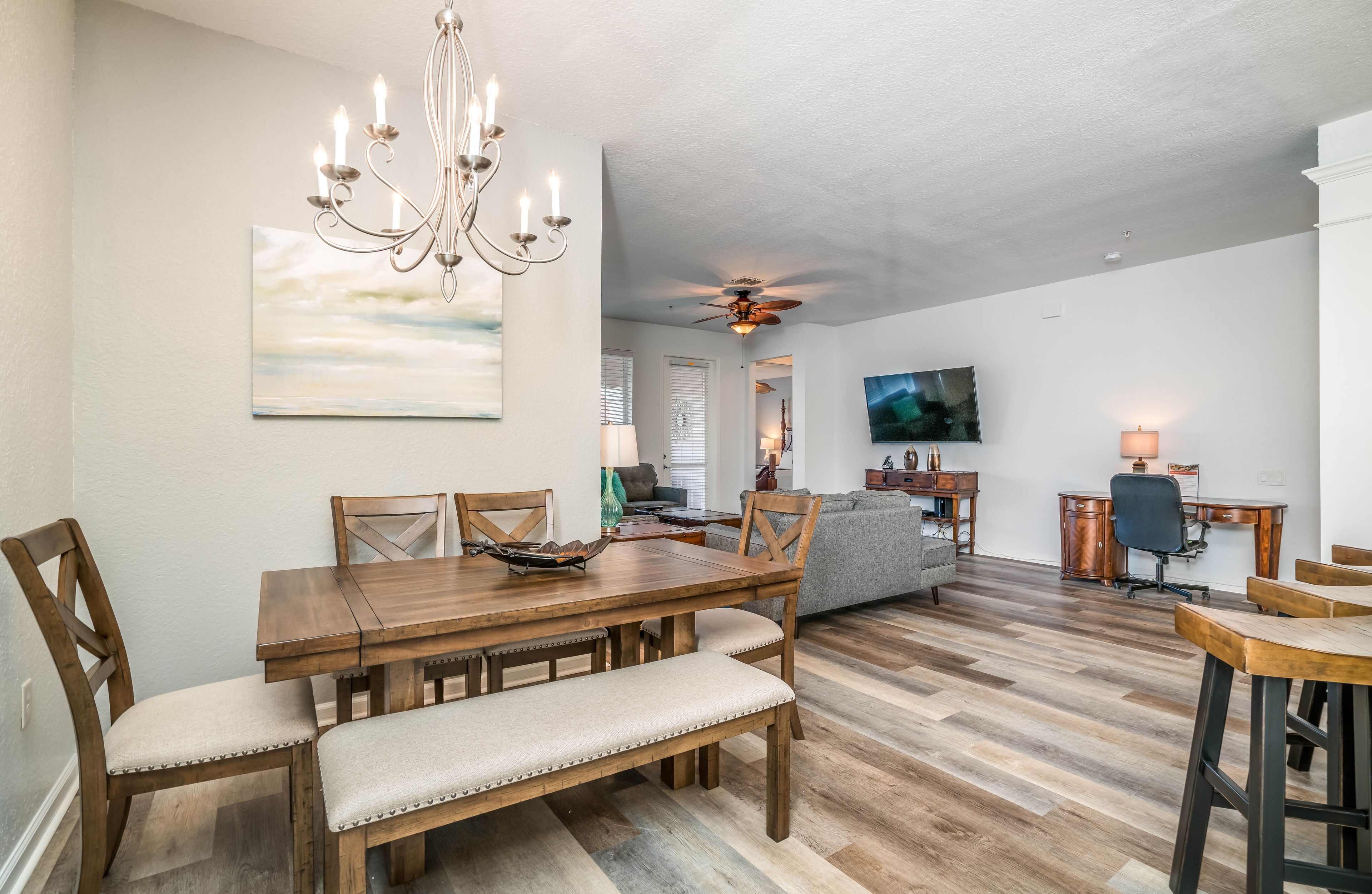 A spacious living area with a wooden dining table, cushioned chairs, a chandelier, a ceiling fan, and a television.
