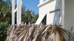 A white building with a wooden porch and a large pile of dried palm leaves in front.