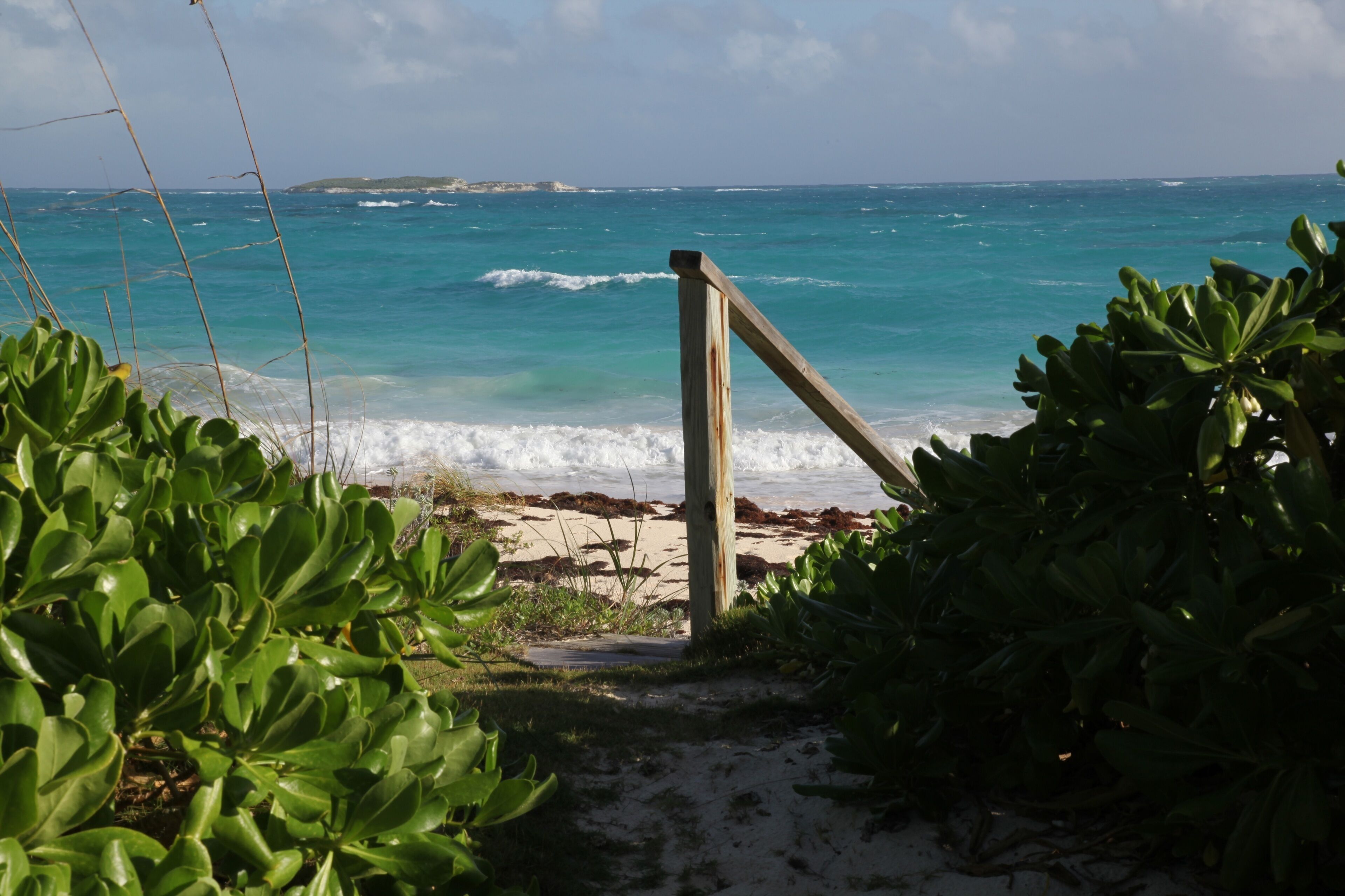 Un sentier en bois mène à une plage bordée d’une eau turquoise et limpide, avec une terre lointaine à l’horizon.