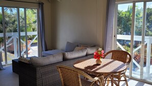 A sunlit room with a high ceiling, a large window, a sofa, a table with chairs, and a ceiling fan.