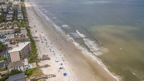 Vista aérea de una playa con una extensa franja de arena, algunos edificios y una clara visión del océano.