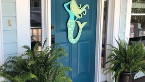 Una puerta azul aguamarina con decoración de sirena, flanqueada por dos helechos en macetas y un felpudo negro.