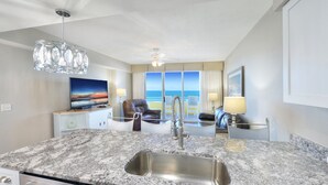 A modern kitchen with a granite countertop, stainless steel sink, and white cabinetry. There is a television on a stand, a ceiling fan, and a view of the ocean through large windows.