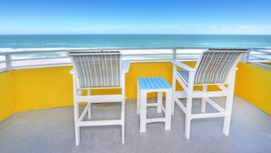 Two white lounge chairs with striped cushions on a balcony with a yellow wall, overlooking the ocean.