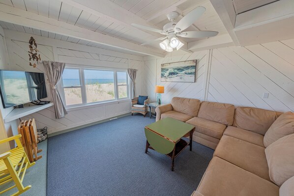 Ein Zimmer mit einem großen Fenster, einem Deckenventilator, einem grünen Tisch, einem beigefarbenen Sofa und einem Fernseher auf einem Schrank.