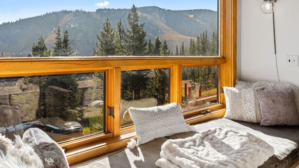 A room with a large window offering a mountain view, a cozy seating area with a blanket and pillows, and a lamp on the wall.