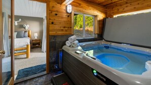 A wooden cabin interior with a hot tub, a bed, and a window overlooking greenery.