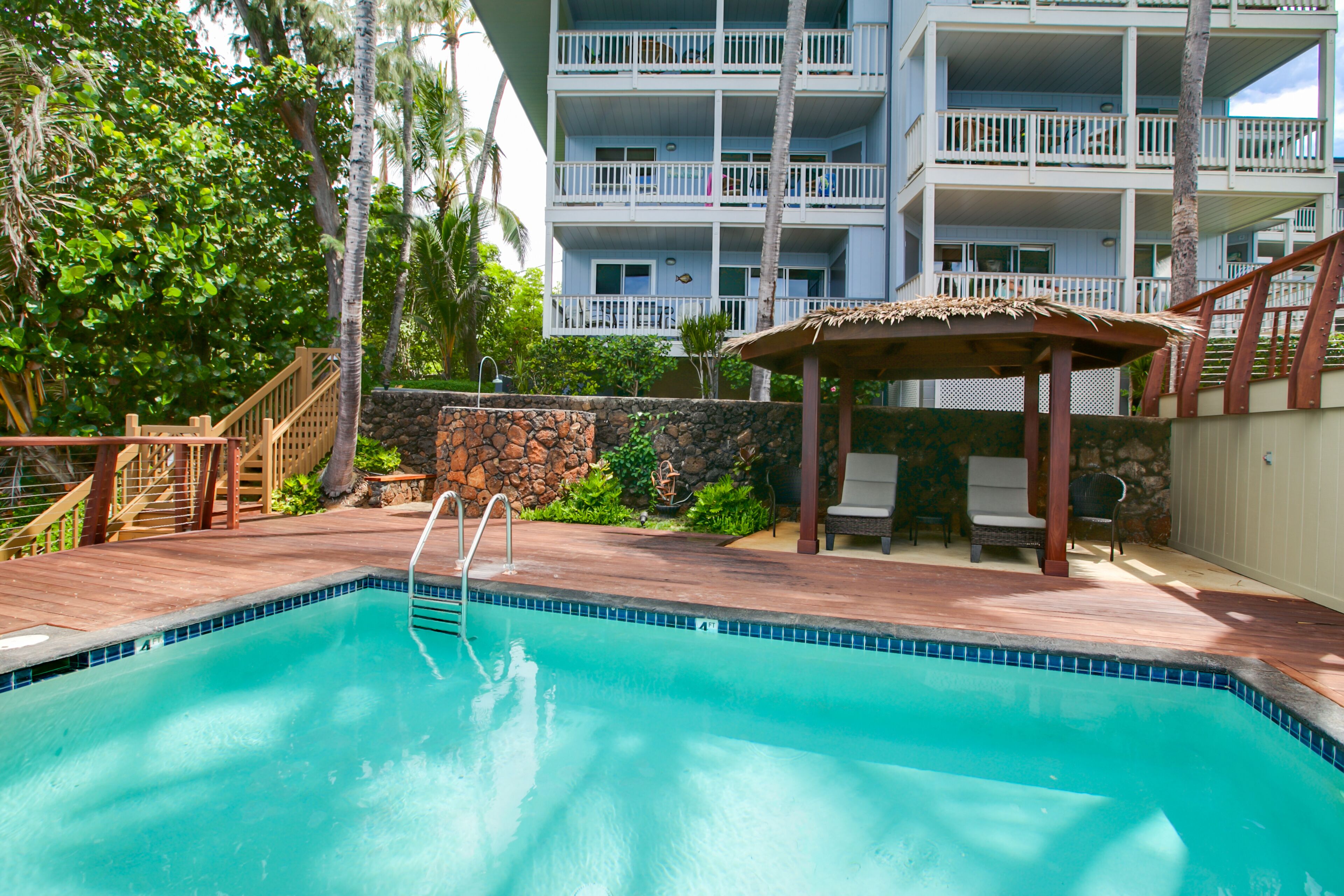 A swimming pool with a wooden deck, a stone wall, and a thatched-roof gazebo.
