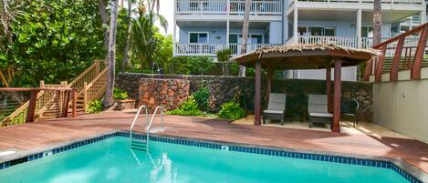 A swimming pool with a wooden deck, a stone wall, and a thatched-roof gazebo.