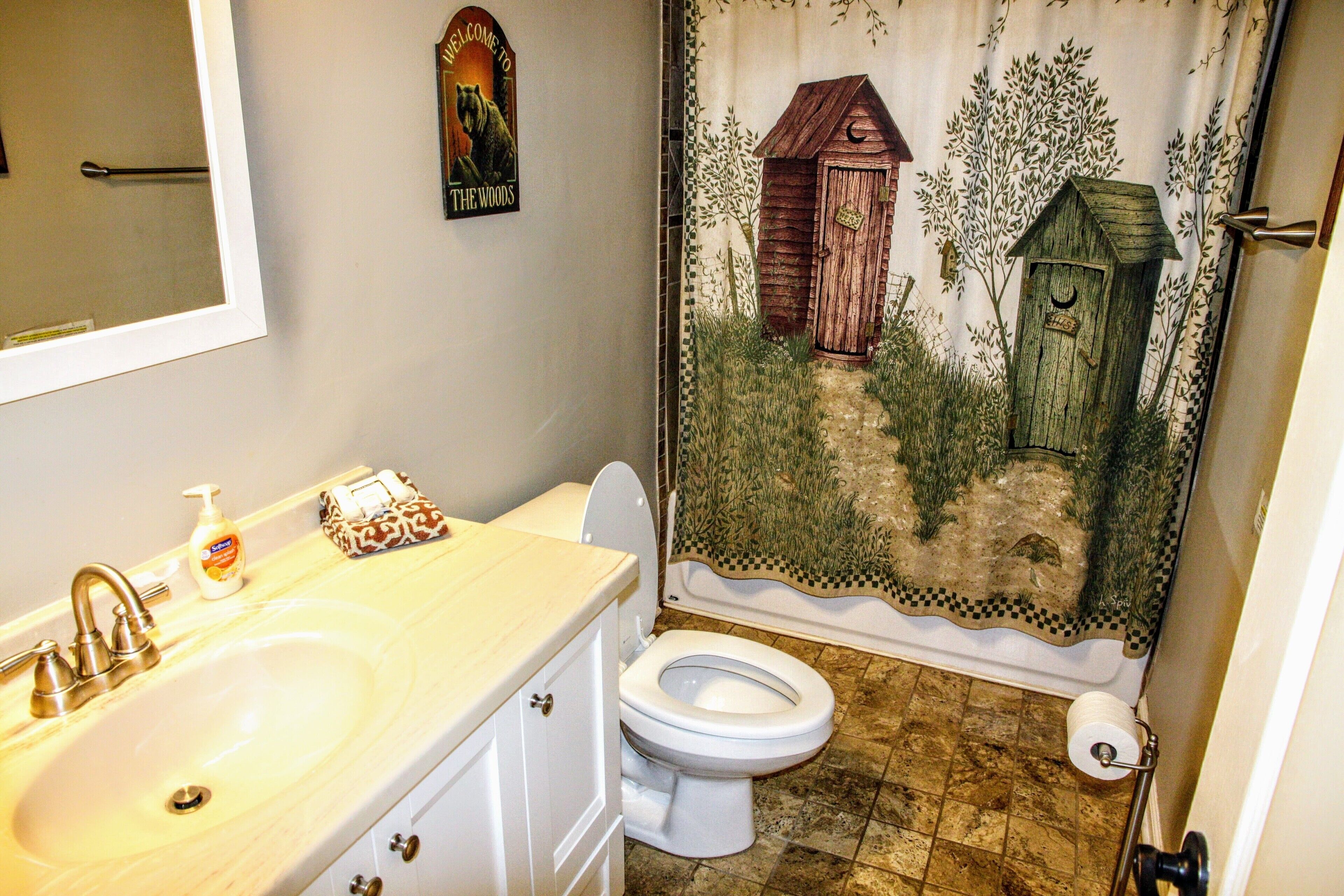 A bathroom with a sink, toilet, and shower curtain depicting a wooden cabin and trees.