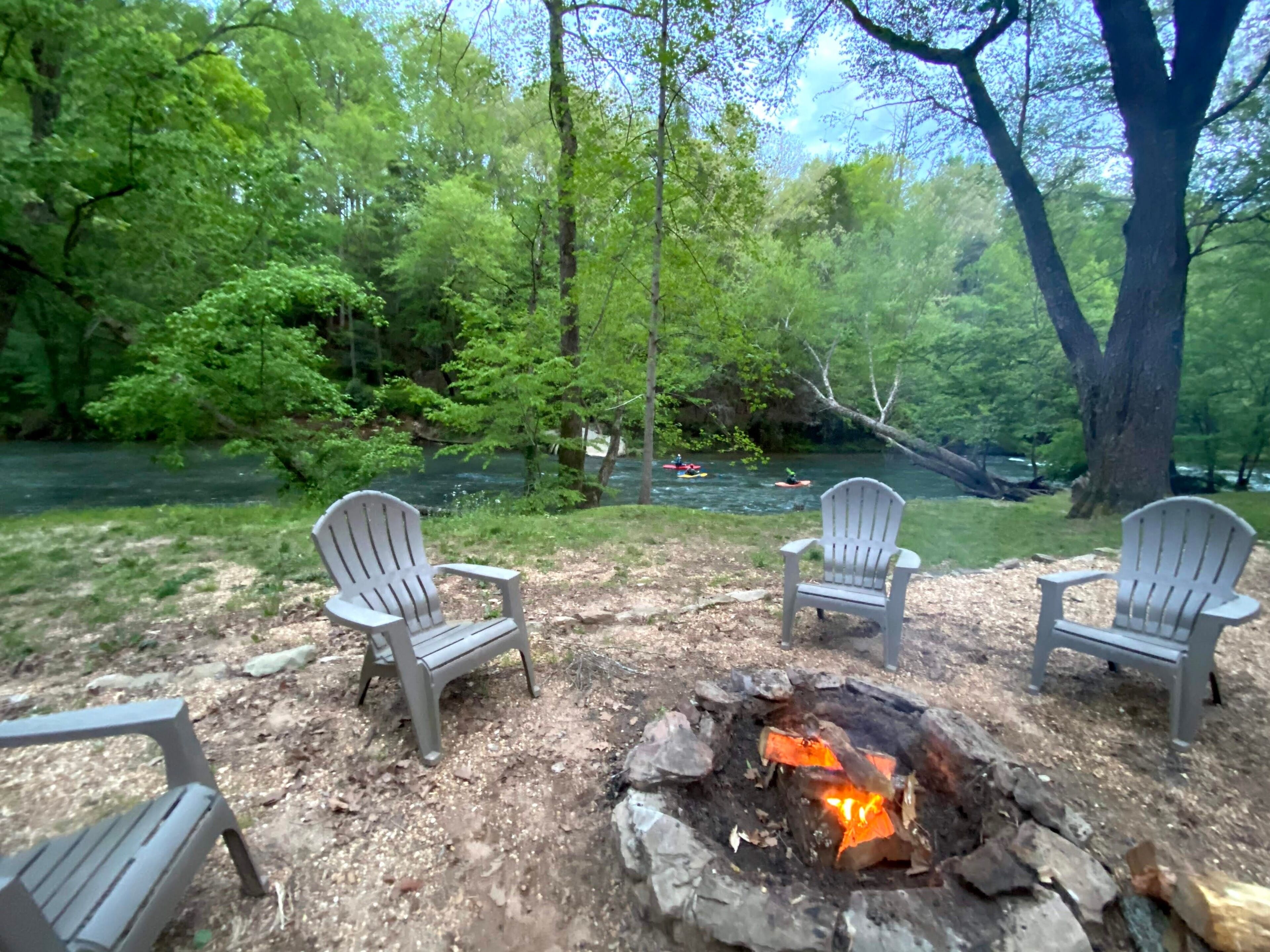 A campsite with three Adirondack chairs, a fire pit, and a river view.