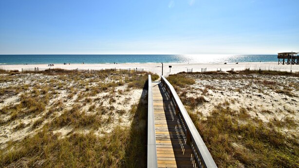 A beach with a wooden boardwalk leading to the water, sandy dunes, and a pier. - Time to get… 🎶 Knee deep in the water somewhere! 🎶 on island time! (Gulf Shores)