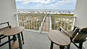 A balcony with a view of the beach, featuring a wooden deck, white railing, and two wooden tables with chairs. - Time to get… 🎶 Knee deep in the water somewhere! 🎶 on island time! (Gulf Shores)