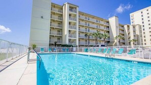 A swimming pool with a white fence, lounge chairs, and a multi-story building with balconies in the background. - Time to get… 🎶 Knee deep in the water somewhere! 🎶 on island time! (Gulf Shores)