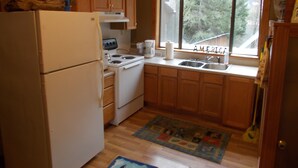Une cuisine de taille réduite, dotée de meubles en bois, d’un réfrigérateur blanc, d’une cuisinière et d’une fenêtre donnant sur les arbres.