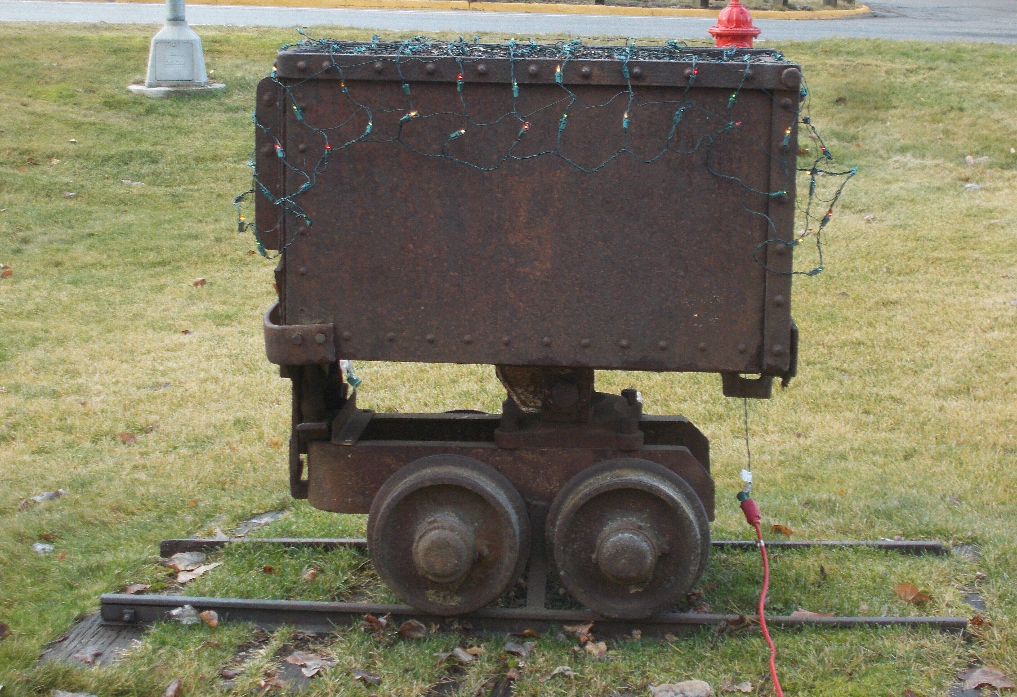 Un carrito de metal oxidado con dos ruedas y un hidrante rojo encima, ubicado en un área cubierta de césped con una carretera al fondo.
