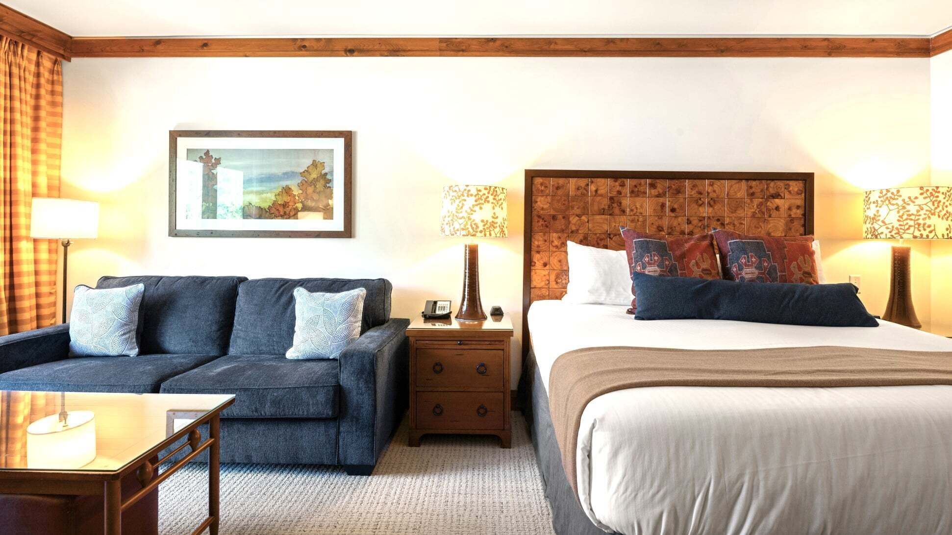 A hotel room with a bed, a sofa, a nightstand, a lamp, and a framed picture on the wall.