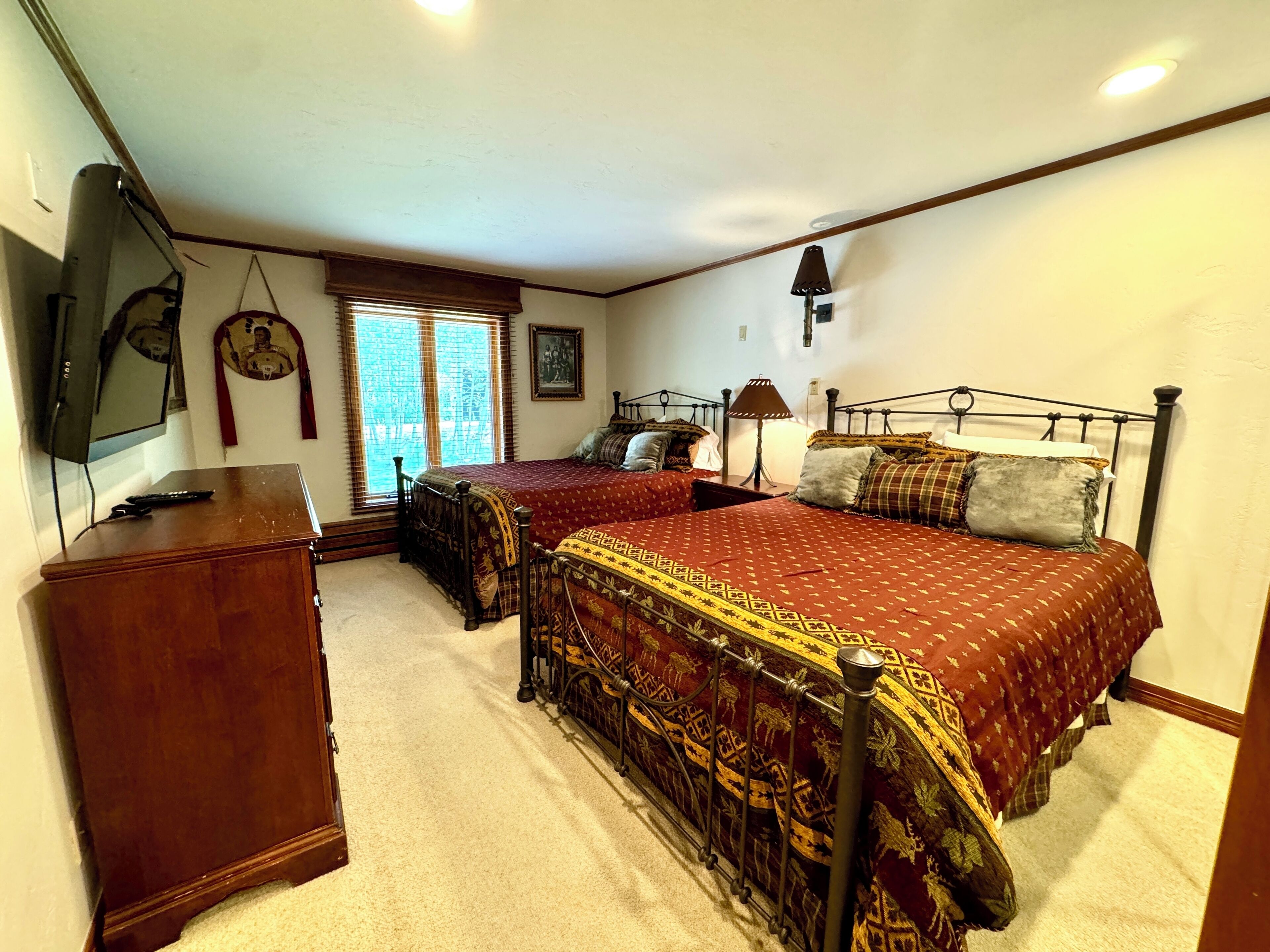 A bedroom with two beds, a dresser, a desk, a lamp, and a window with blinds.