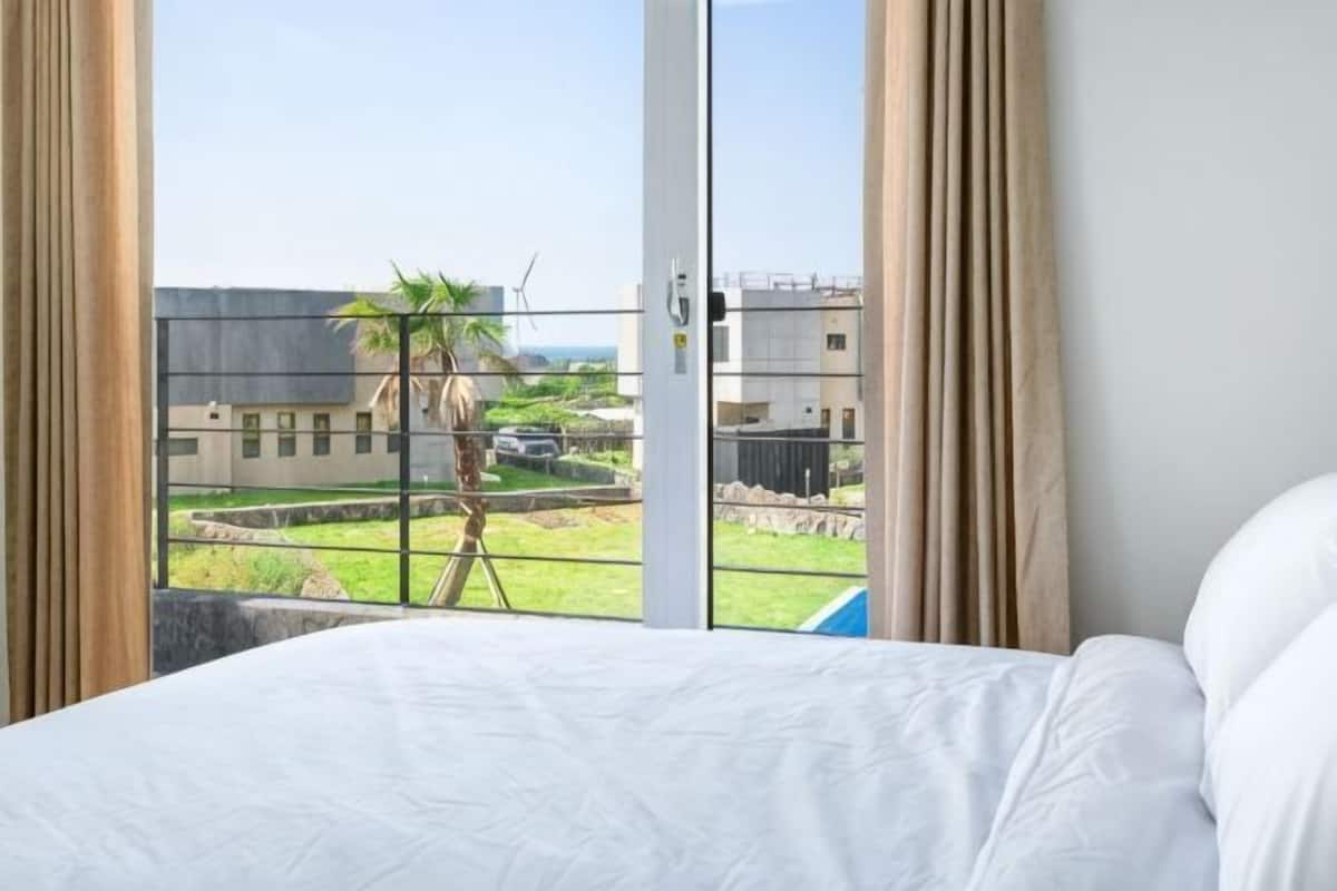Quarto de hotel com cama, vista para um edifício e uma palmeira, e varanda com guarda-corpo.