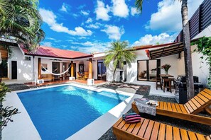 Zona junto a la piscina con una pileta rectangular, sillas de lounge de madera, una hamaca y una parrilla.