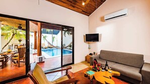A modern living room with a grey sofa, wooden dining table, and chairs, a TV mounted on the wall, and a pool visible through the glass doors. - Casa Tropical☙Remodeled Beach Oasis Perfect for resting and relaxing ❧ (Tamarindo)